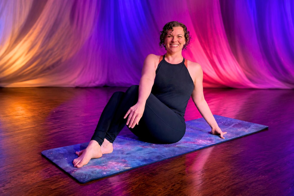Brittani seated on a starry Pilates mat, smiling, stage curtains lit in purple and gold behind her