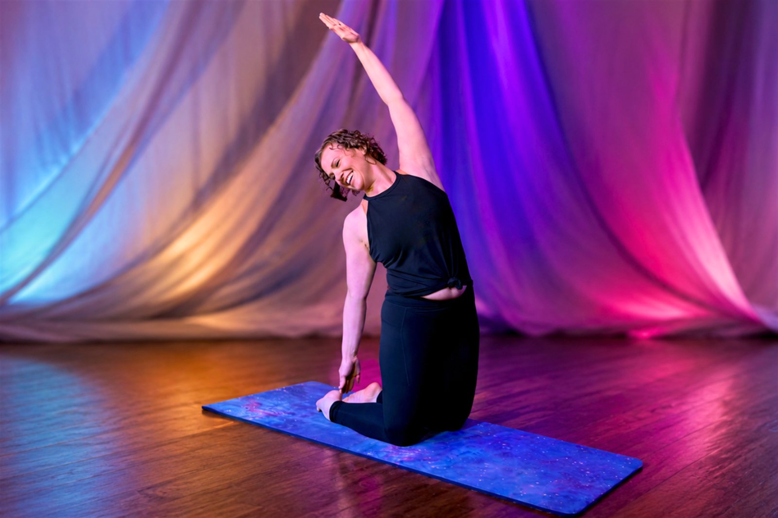 Brittani leading a mat Pilates stretch on a starry mat, stage curtains glowing behind her